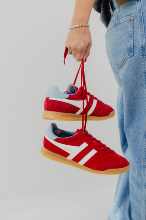 Red sneakers with white accents held by a person wearing blue jeans against a light gray background