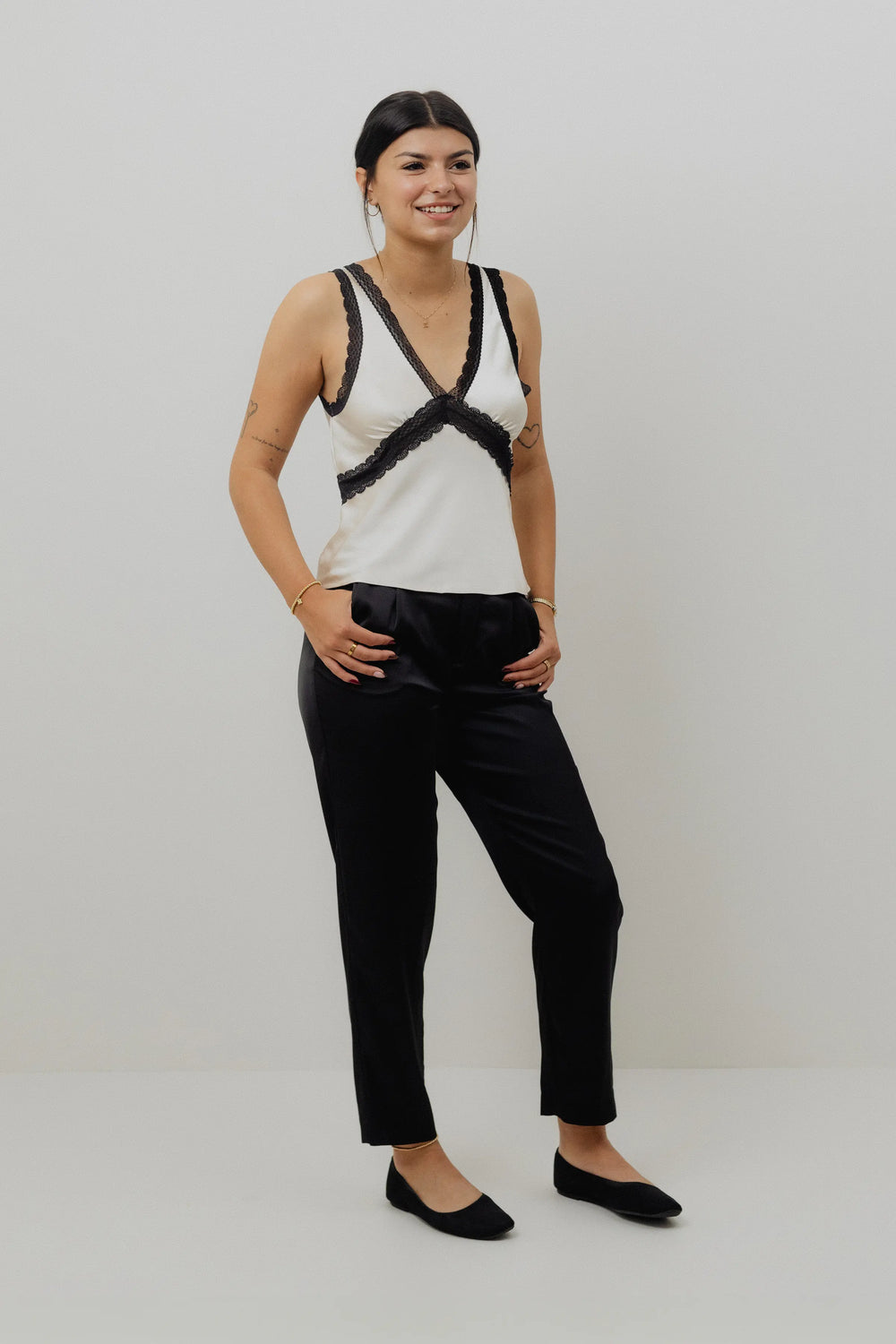 Woman wearing a white tank top with black lace details and black pants on a white background