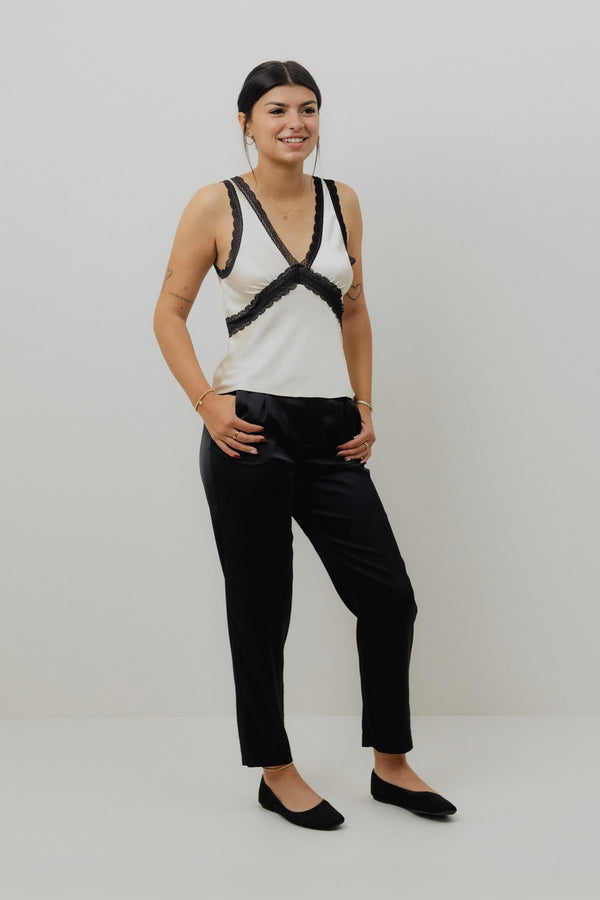 Woman wearing a white tank top with black lace details and black pants on a white background