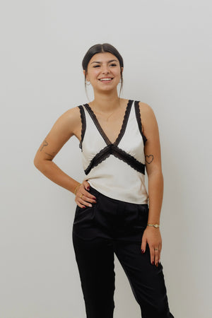Woman wearing a white sleeveless top with black lace details and black pants against a plain background