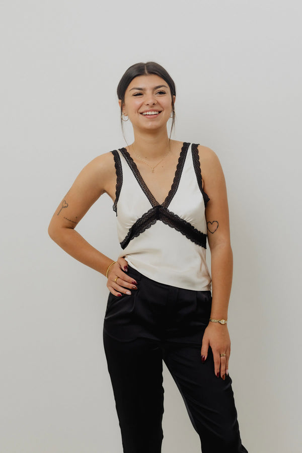 Woman wearing a white sleeveless top with black lace details and black pants against a plain background