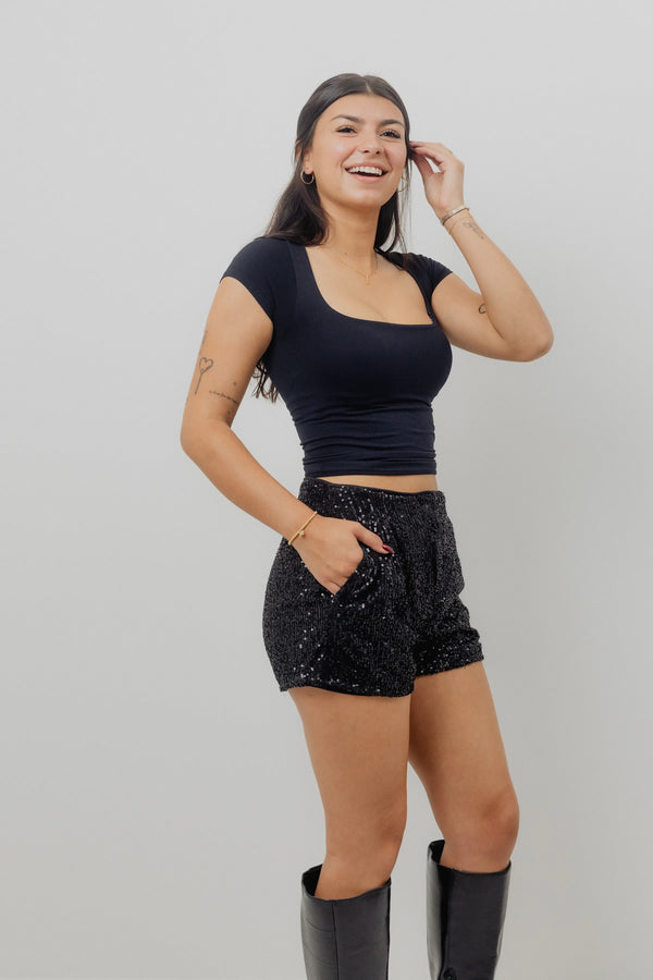 Woman wearing a black crop top and black sequin shorts on a plain background