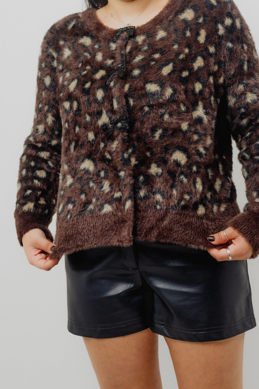 Person wearing a brown patterned sweater, black leather shorts, and black knee-high boots on a light gray background