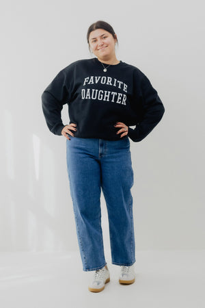 Person wearing a black sweatshirt with 'FAVORITE DAUGHTER' text and blue jeans on a white background