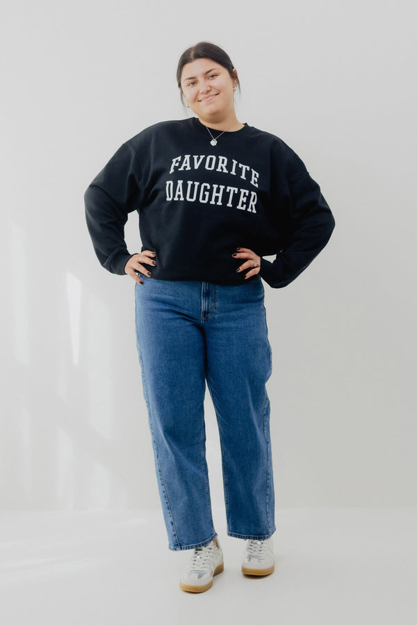 Person wearing a black sweatshirt with 'FAVORITE DAUGHTER' text and blue jeans on a white background