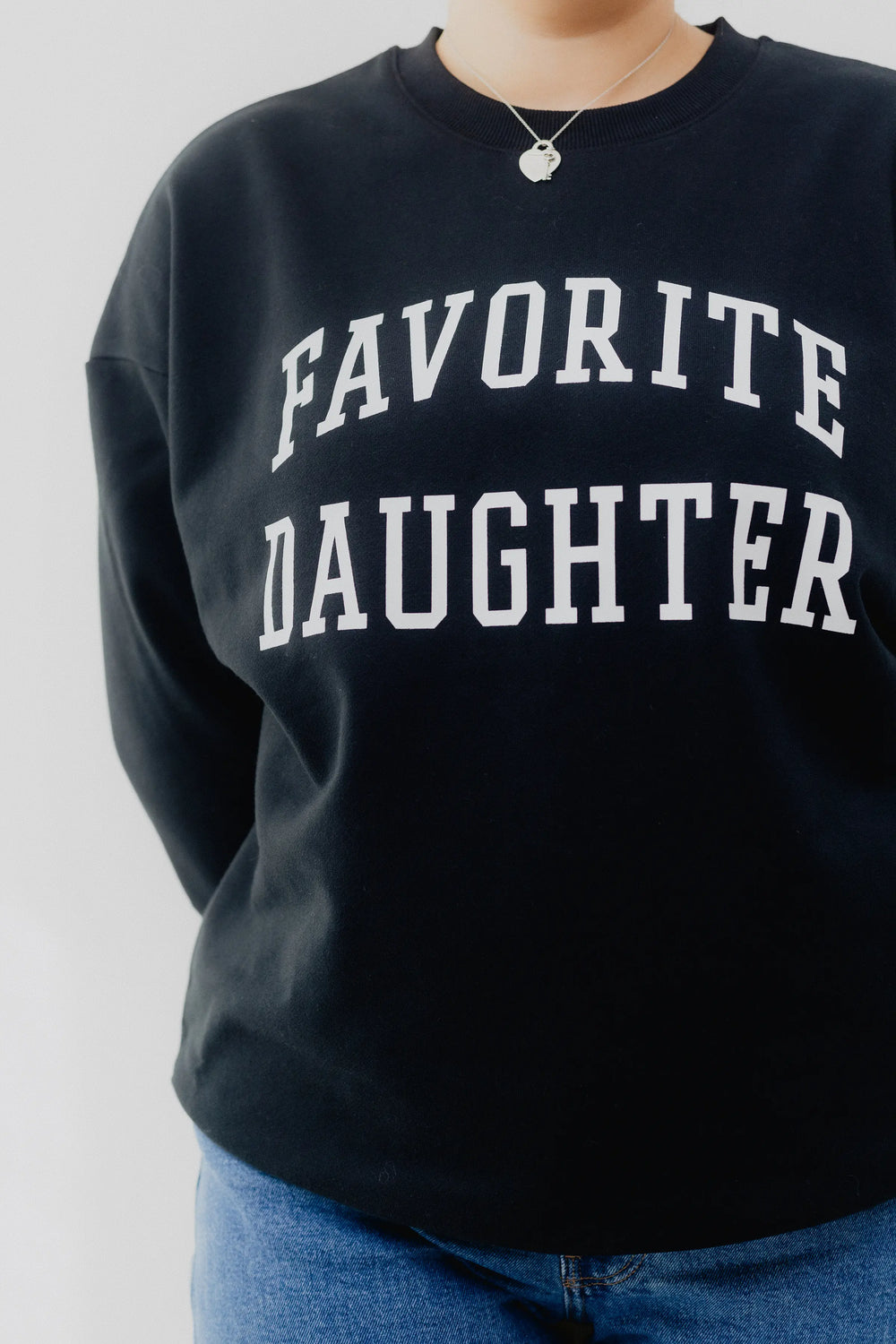 Person wearing a black sweatshirt with 'FAVORITE DAUGHTER' text and blue jeans on a white background